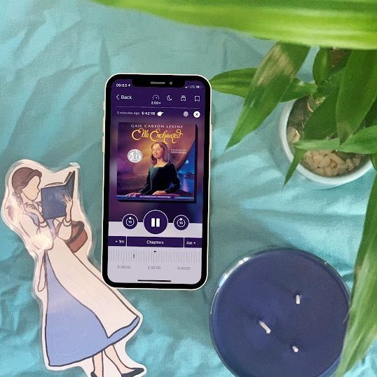 Katie's iPhone, showing the audiobook of "Ella Enchanted" by Gail Carson Levine is on a blue background and surrounded by a variety of items - including a purple candle, a bamboo plant, and a Belle (Disney Princess from "Beauty and the Beast") bookmark.